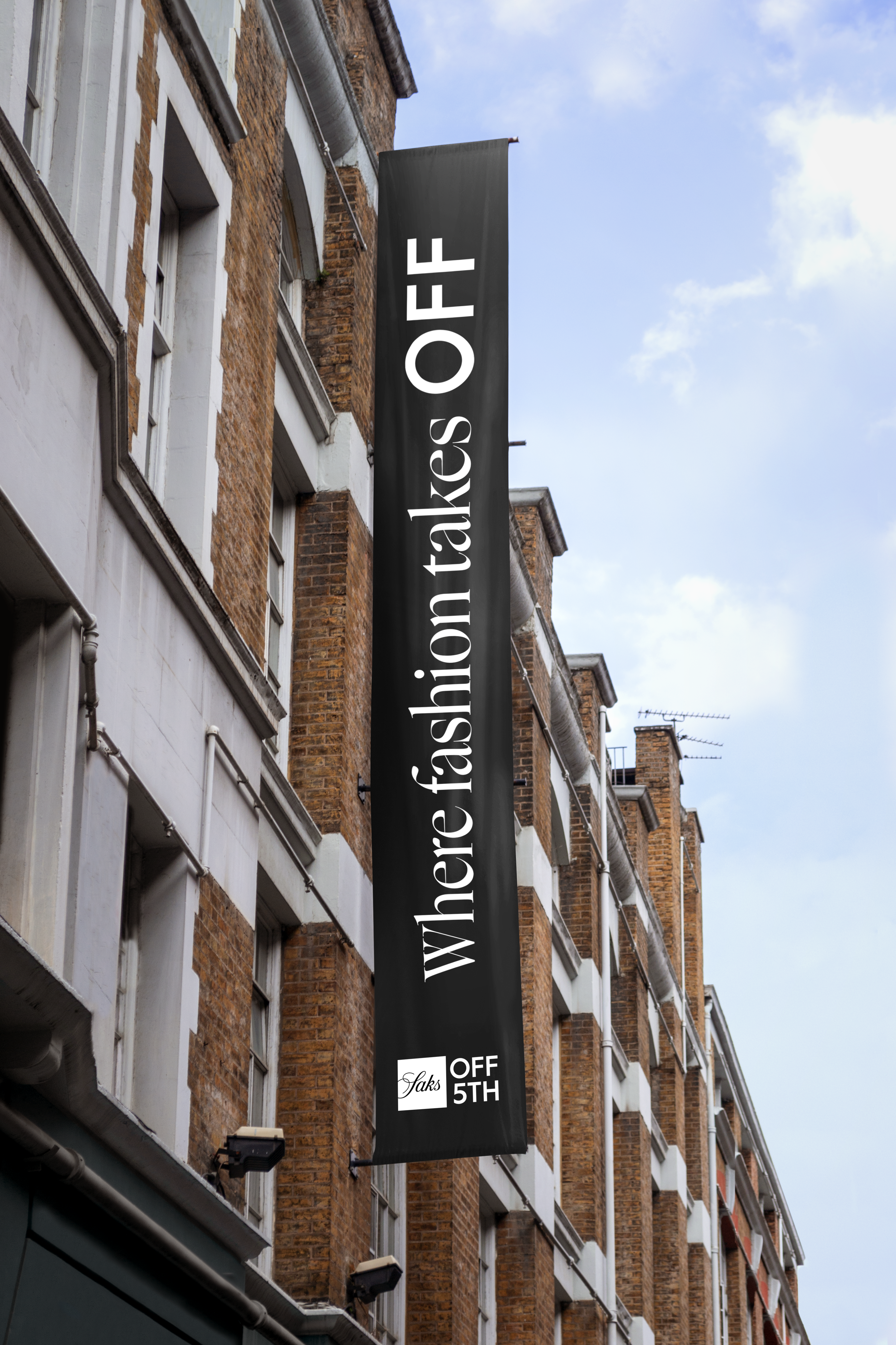 Saks OFF 5TH banner in black saying "Where fashion takes OFF" in white and the logo at the bottom. The banner is next to a brick building in front of blue sky.