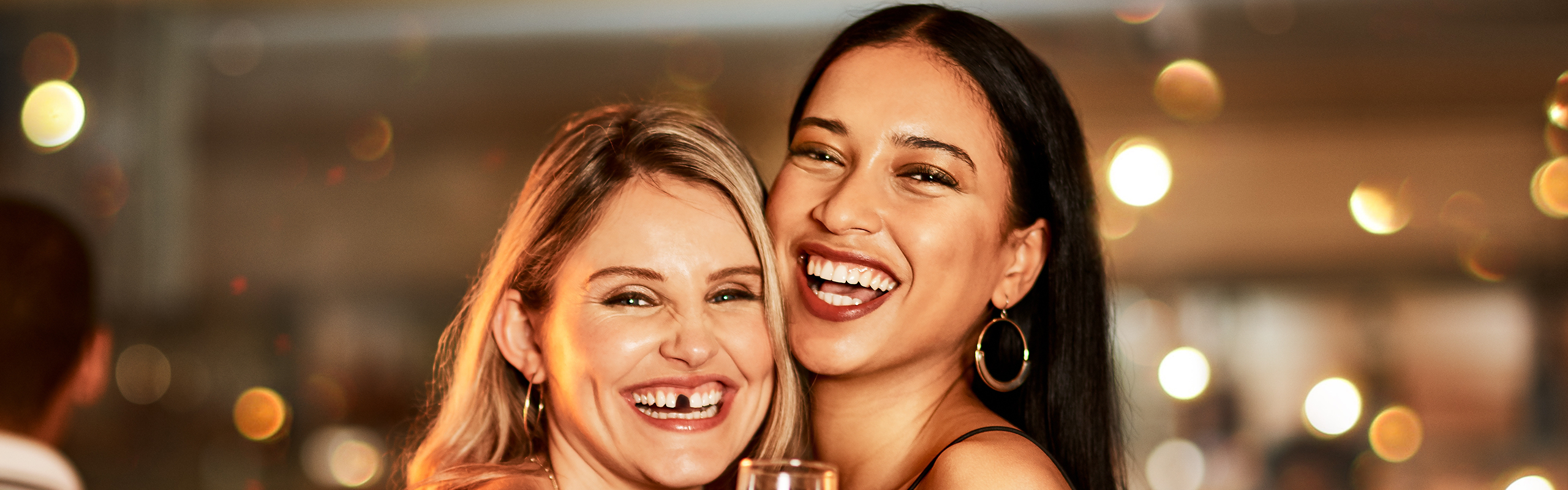 zwei Freundinnnen lachend mit einer Zahnlücke in einer Bar - Zahnzusatz-Experten, sichern Sie sich Zahnbehandlungen von Experten mit unseren Zahnzusatzversicherungen 