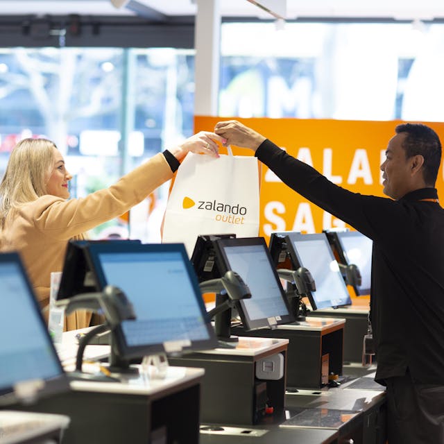 A person behind a checkout hands a customer a paper bag with the Zalando Outlet logo on it.