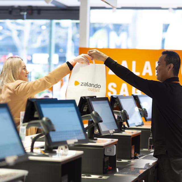 A person behind a checkout hands a customer a paper bag with the Zalando Outlet logo on it. 