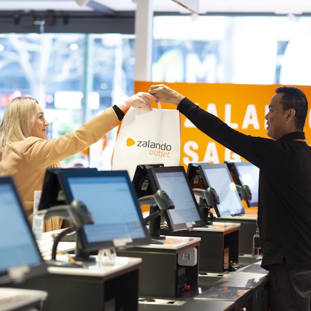 A person behind a checkout hands a customer a paper bag with the Zalando Outlet logo on it. 