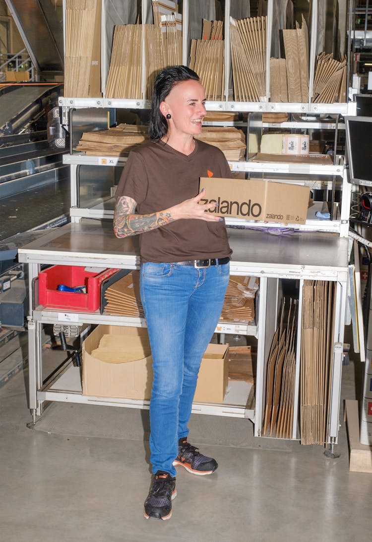 A person standing while holding a package box in their hands and a big pile of packages behind them