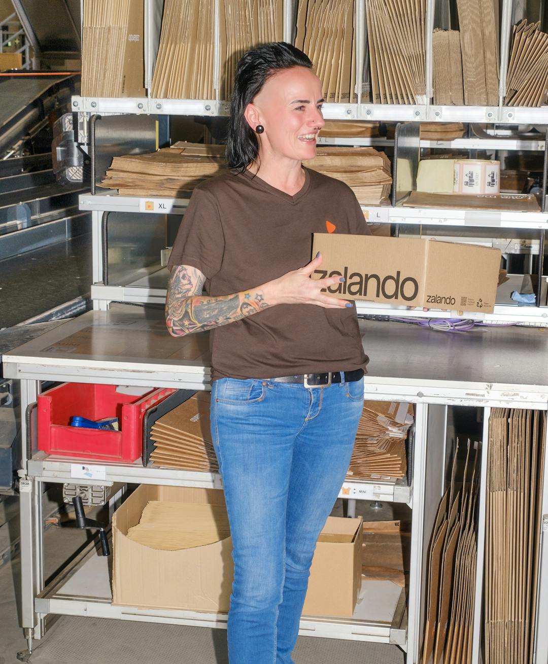 A person standing while holding a package box in their hands and a big pile of packages behind them