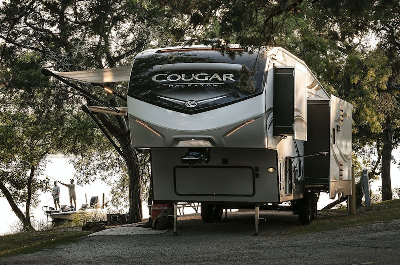 Cougar RV parked in front of a lake with two men fishing on a boat next to it.