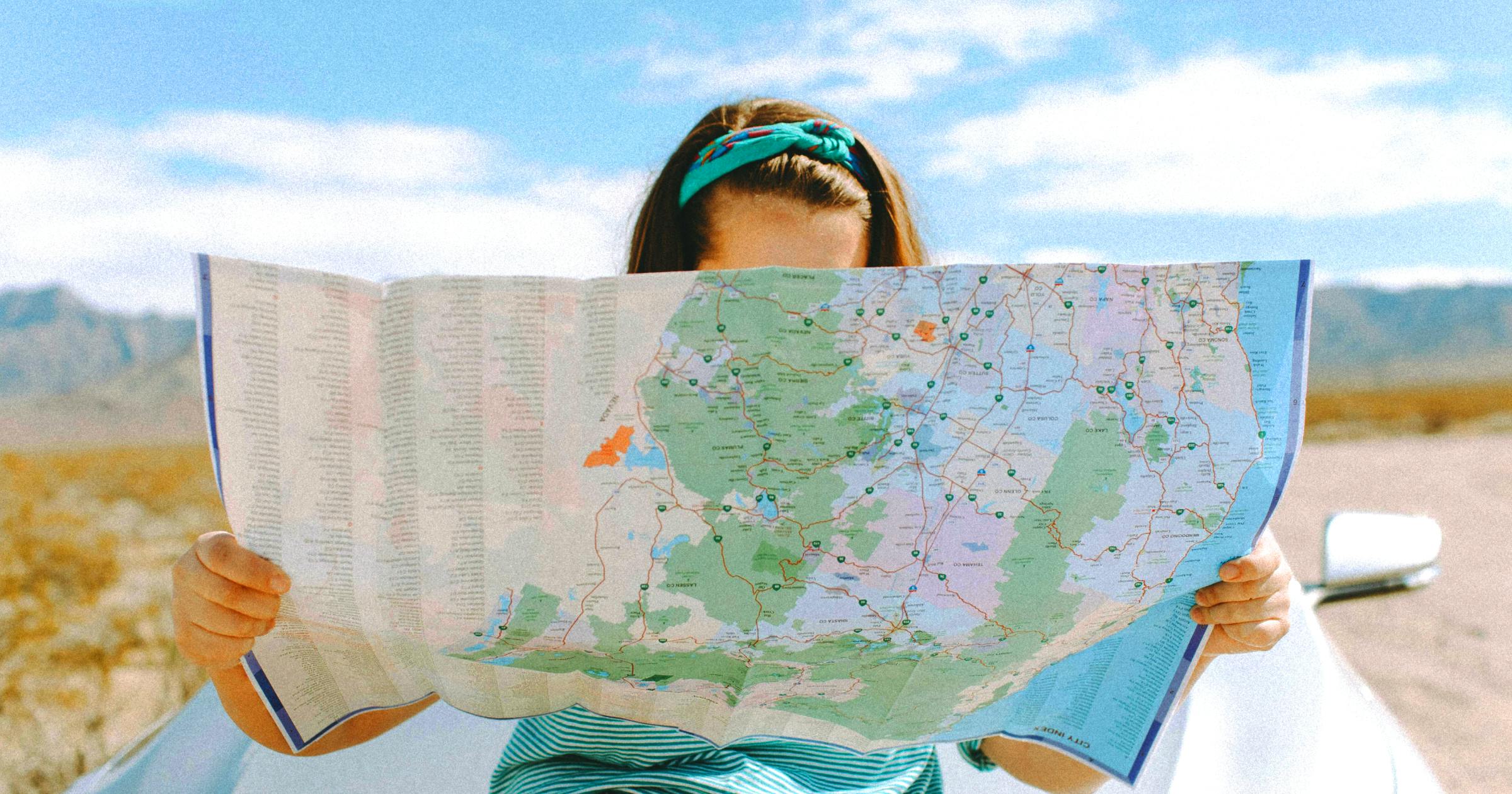 Girl with a blue shirt and headband looking at a map with a car and landscape behind her