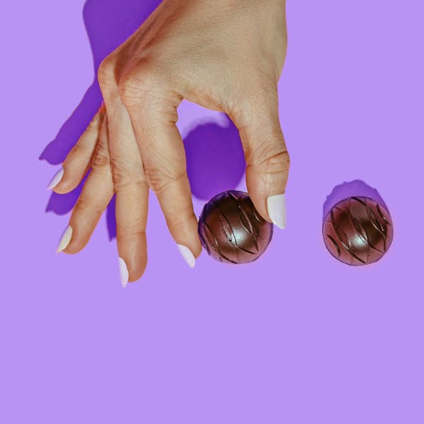 Woman with white finger nail polish holding a chocolate truffle with a purple background