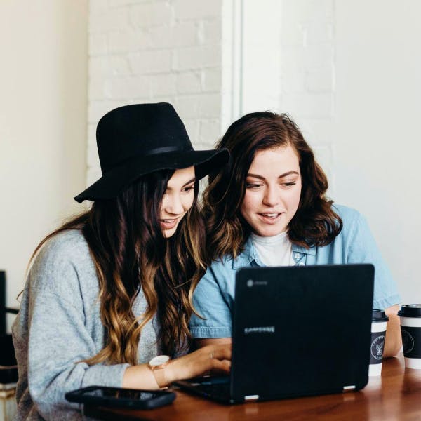 Two young brunette women booking a trip on a computer at a cafe with coffees on the table