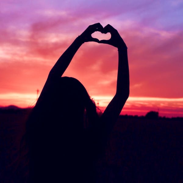 Girl making a heart with her hands with a pink sky in the background