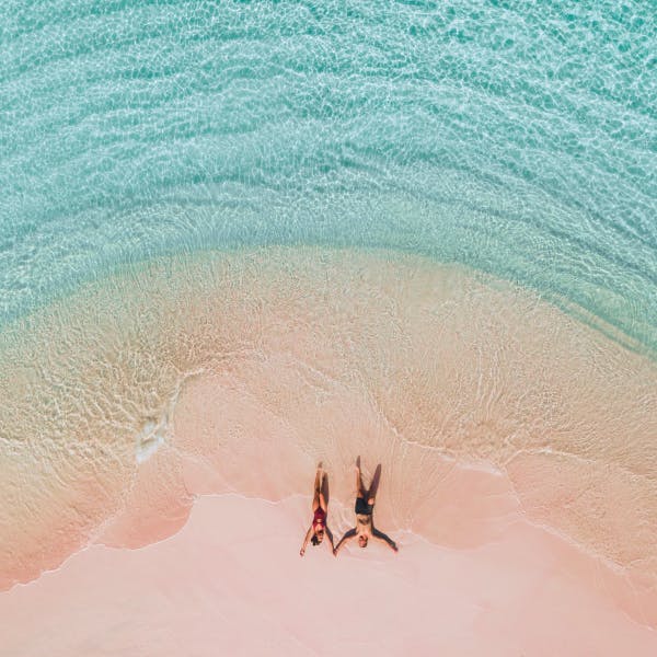 A couple laying in their bathing suits on a pink sand beach surrounded by turquoise water