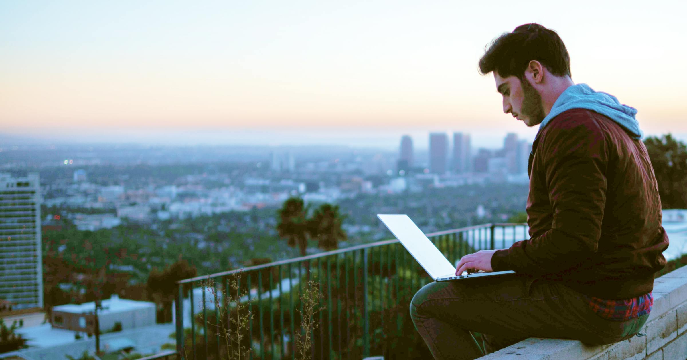Man sitting on balcony working on a laptop with city view in the background