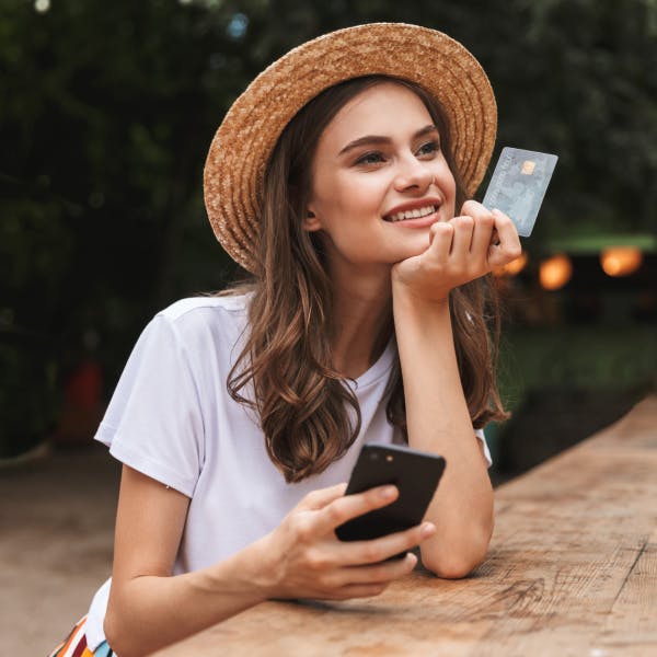 A girl with a white shirt and straw hat holding a credit card and her phone smiling