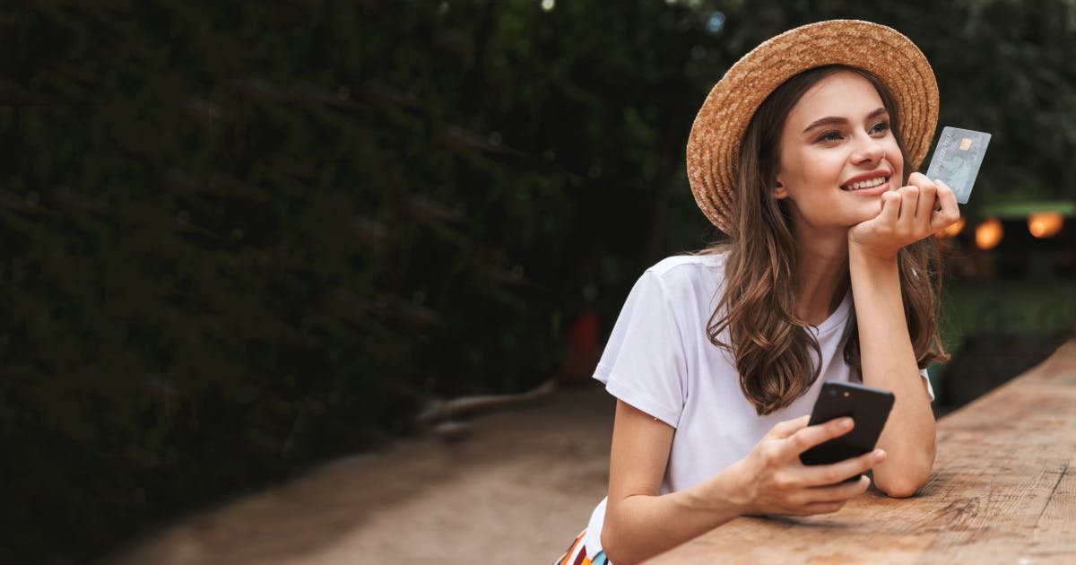 A girl with a white shirt and straw hat holding a credit card and her phone smiling