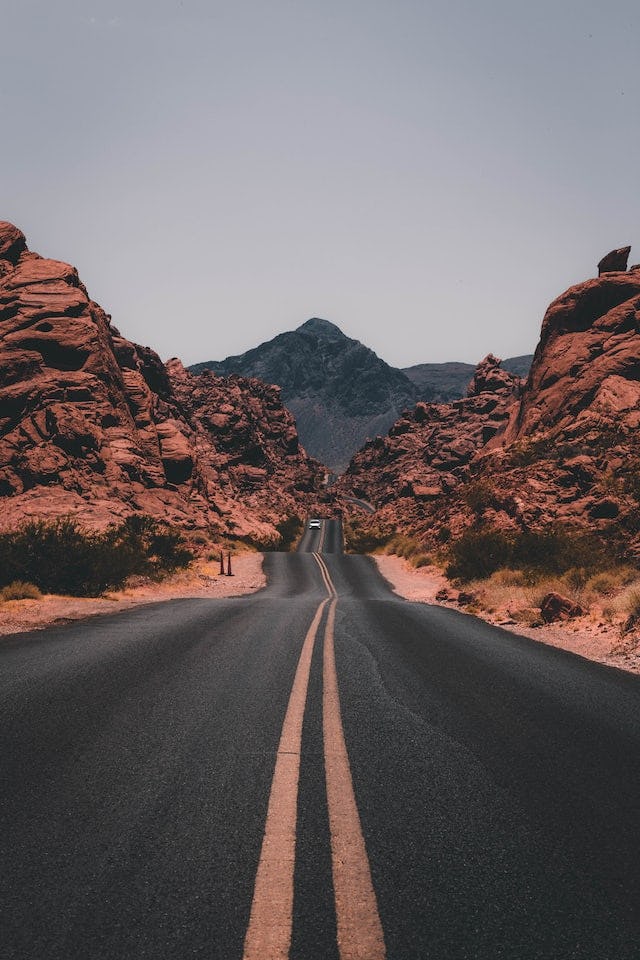 Paved road with red rocks around and a mountain in the bg with one car coming towards the camera