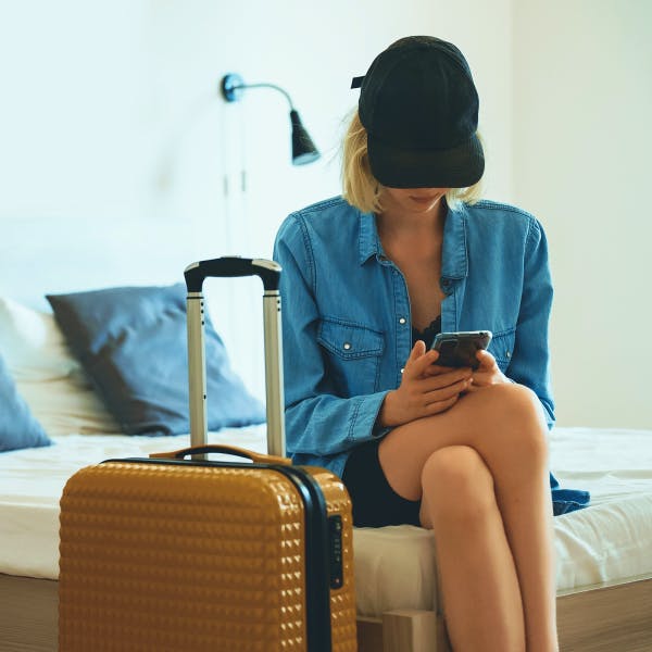Woman sitting on a bed in a black hat and a deni shirt with luggage next to her checking her phone
