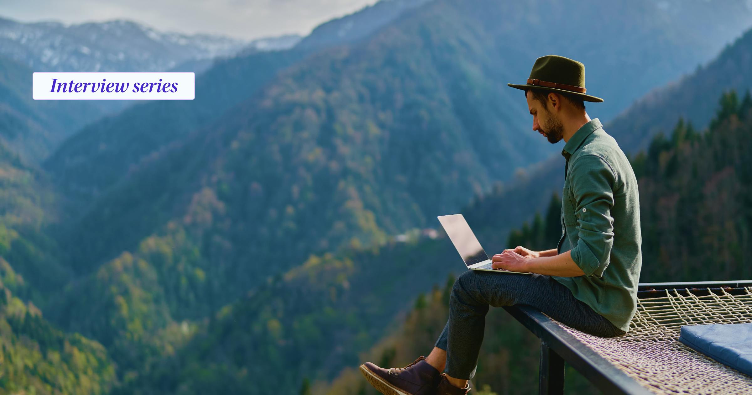 Man with hat sitting on a net with mountains in the background working on his laptop and the words interview series on the image
