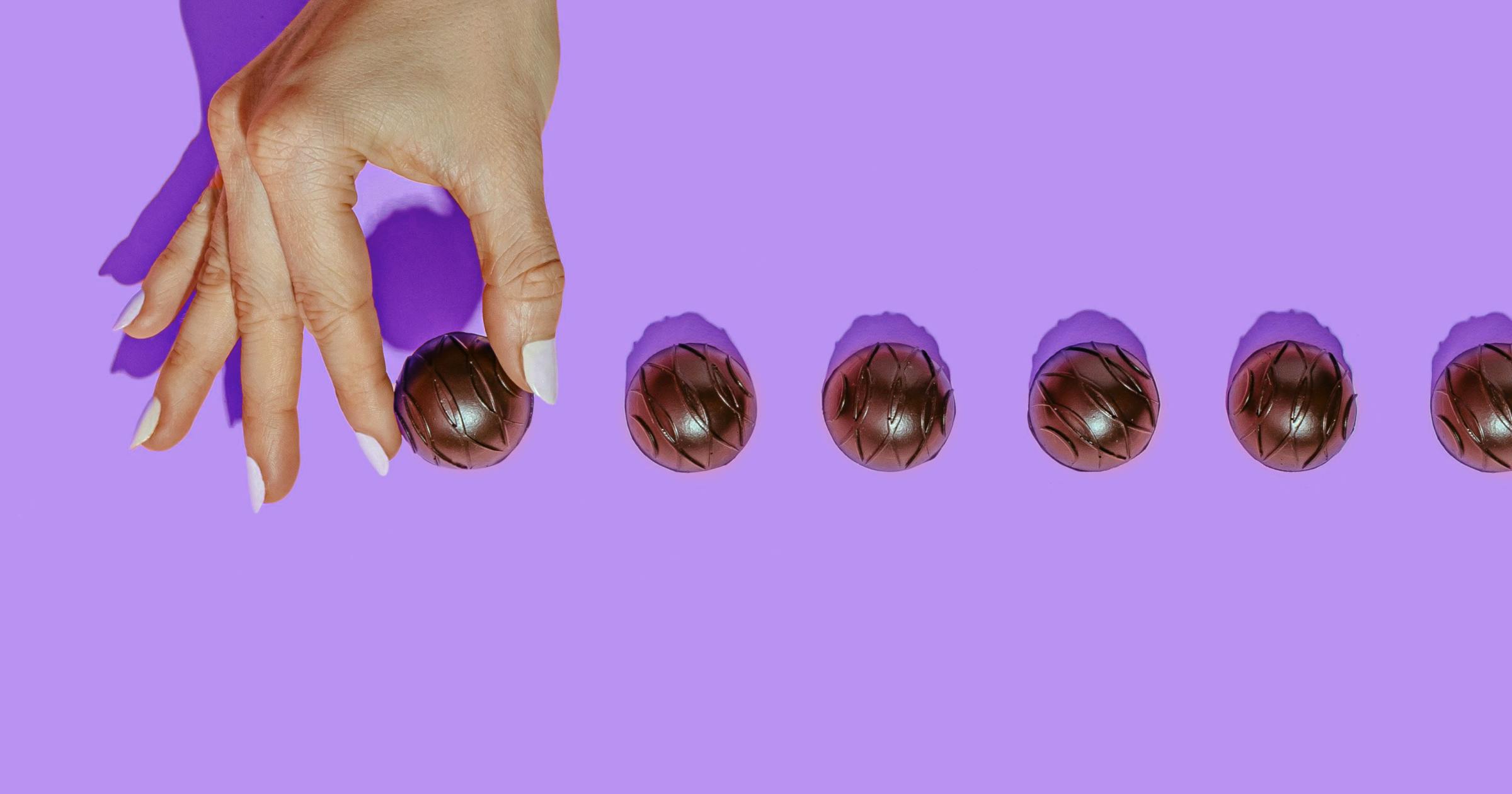 Woman with white fingernail polish holding a chocolate truffle and other chocolate truffles lined up next to it with a purple background