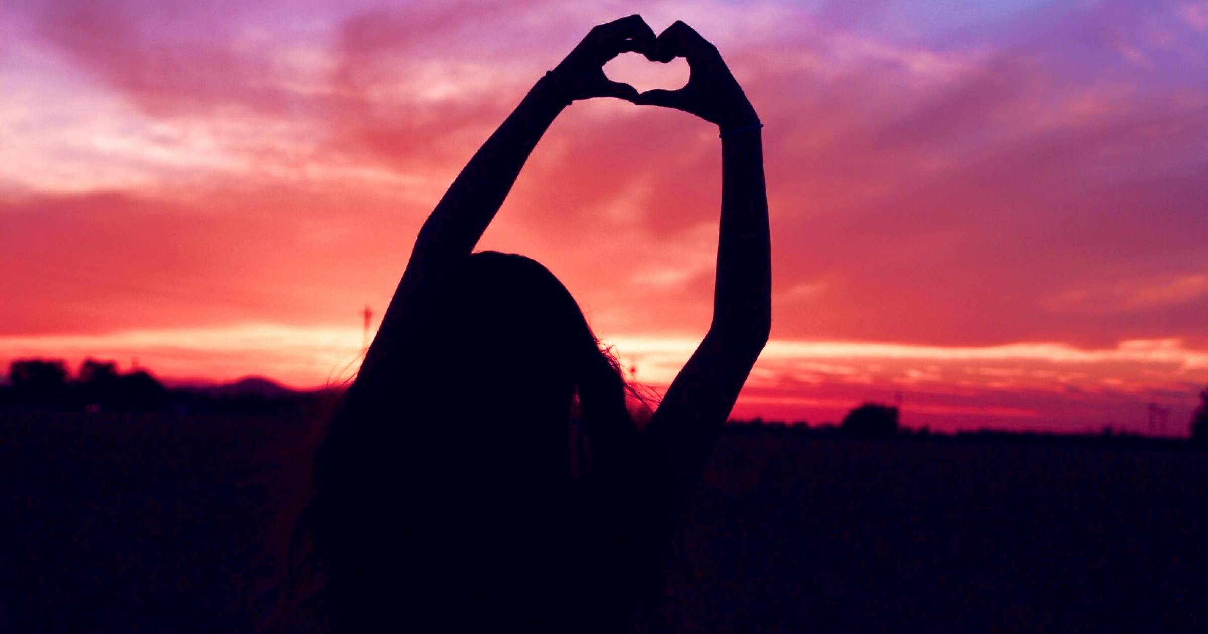 Girl making a heart with her hands with a pink sky in the background