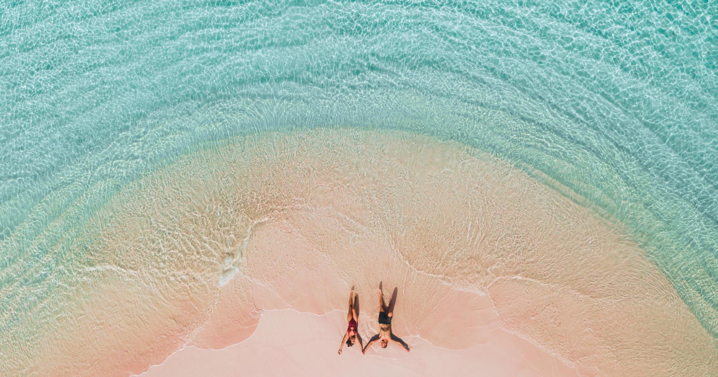 A couple laying in their bathing suits on a pink sand beach surrounded by turquoise water