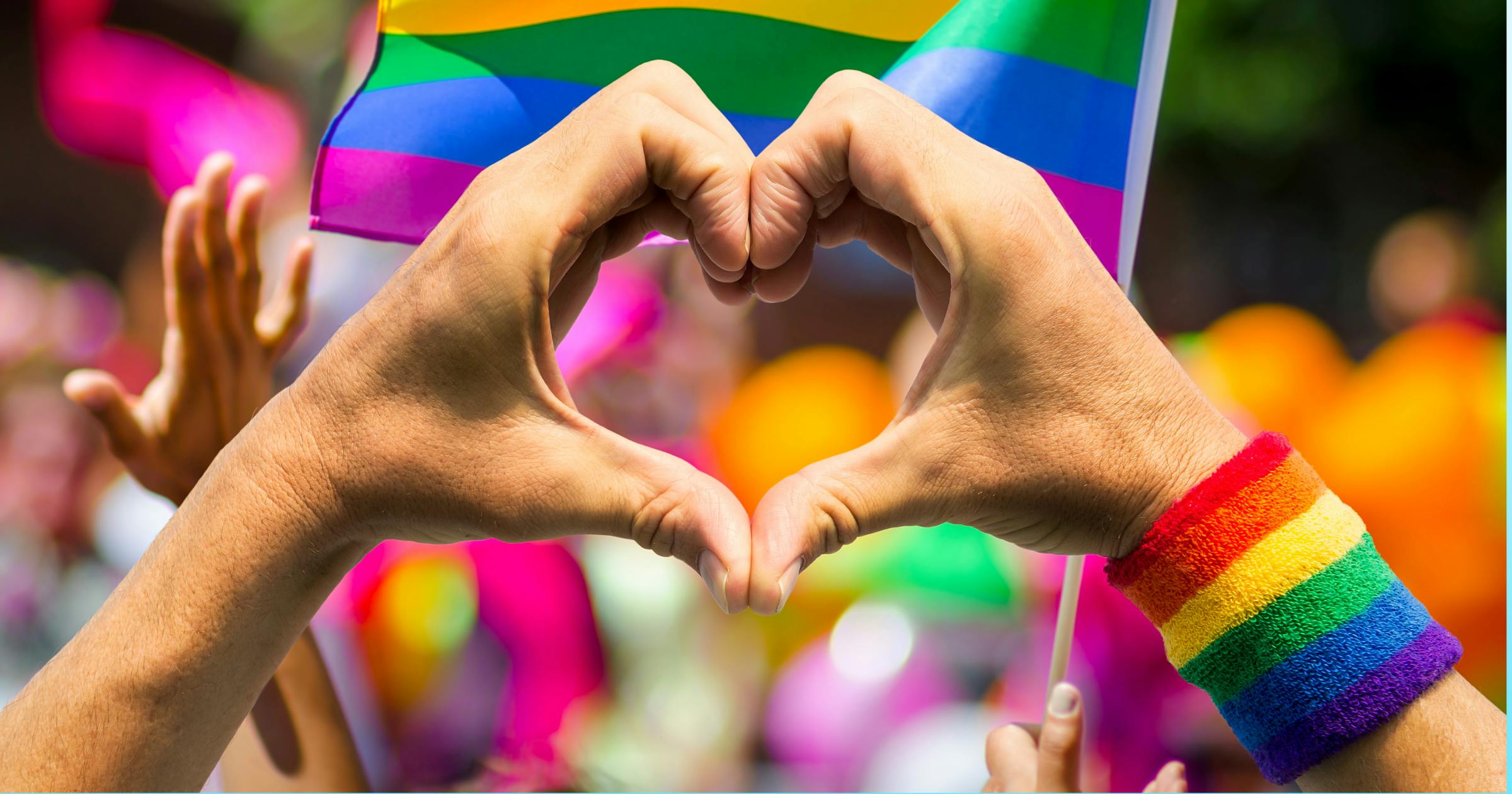 Person making a heart with their hands with a pride celebration in the background.