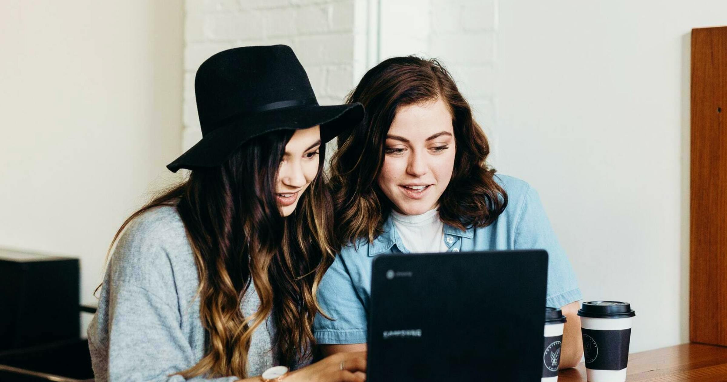 Two young brunette women booking a trip on a computer at a cafe with coffees on the table