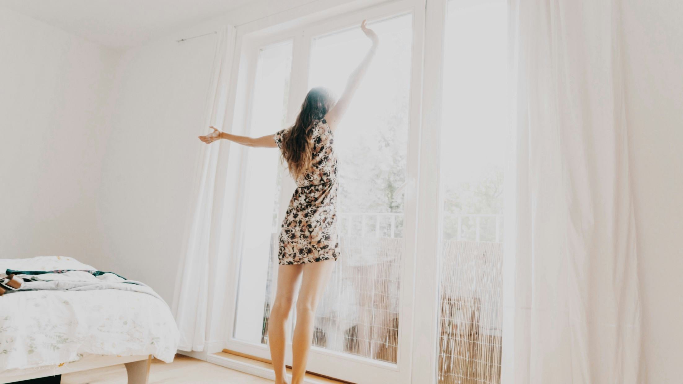 woman in patterned dress stretching in her bedroom looking outside onto her balcony
