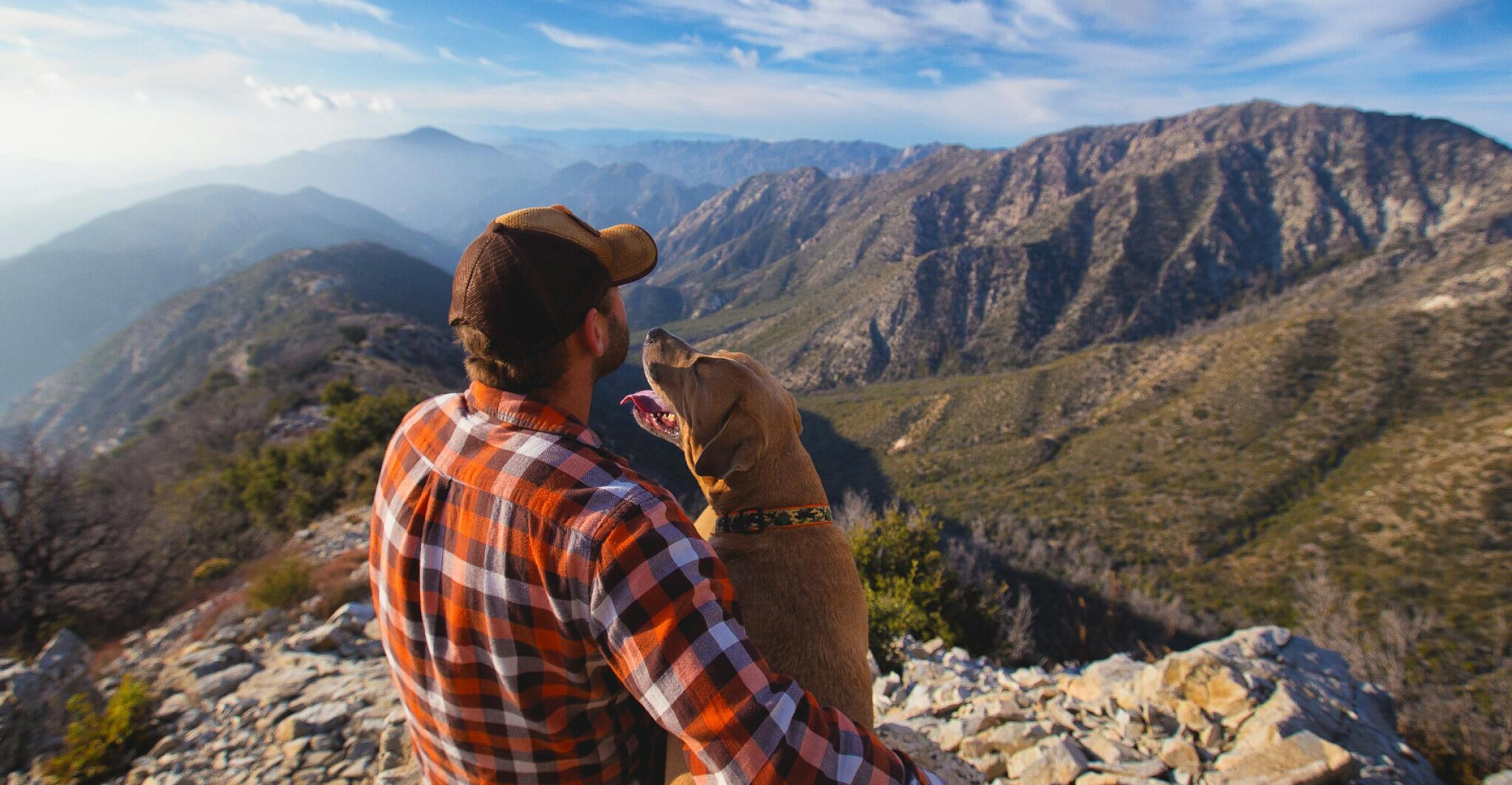 Man in a hat and plaid shirt sitting with his dog overlooking the mountains
