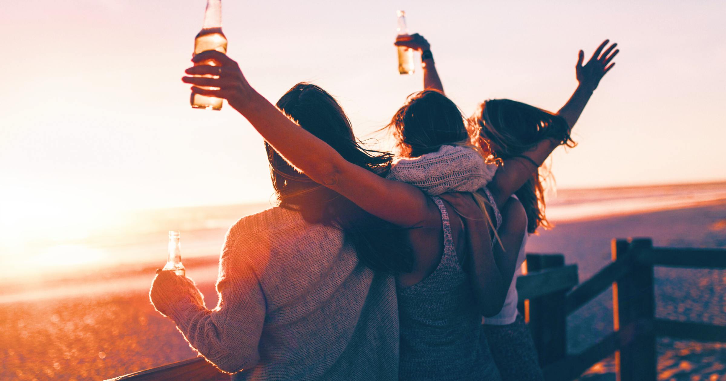 3 girls hugging and raising their arms as they watch the sunset together with beers 