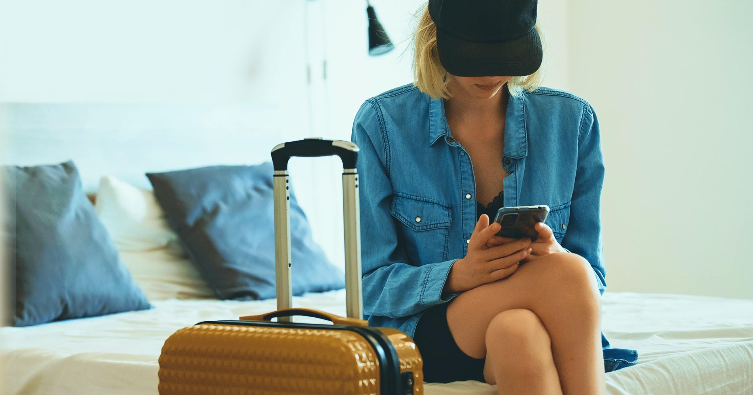Woman sitting on a bed in a black hat and a deni shirt with luggage next to her checking her phone